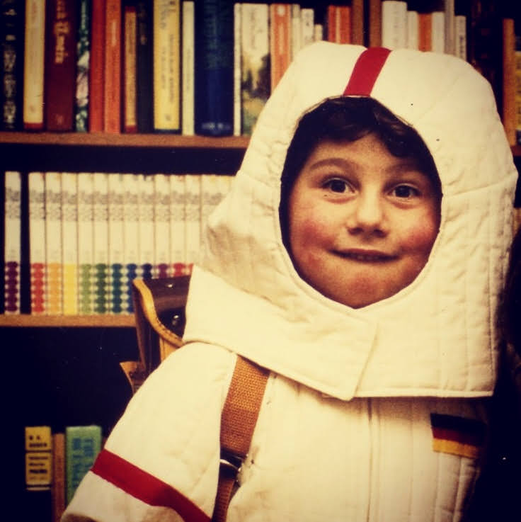 Bernhard Goodwin als Kind in Astronautenverkleidung vor einer Bücherwand.